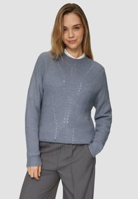 Pull en maille bleu clair avec un col rond, présentant des motifs texturés et de petites embellissements, associé à un pantalon gris.