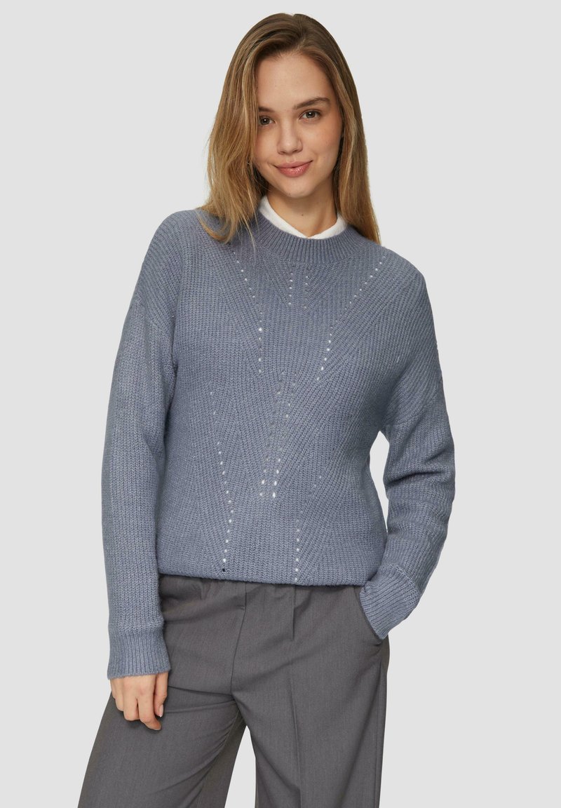 Pull en maille bleu clair avec un col rond, présentant des motifs texturés et de petites embellissements, associé à un pantalon gris.