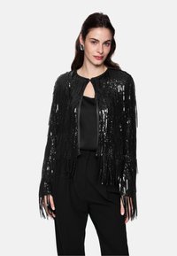 Black sequined jacket with fringe detailing, round neckline, and keyhole front. Worn over a black top and paired with black trousers.