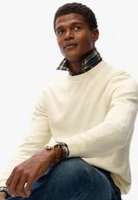 Cremfarbener Pullover mit Rippbündchen über einem karierten blau-grünen Hemd mit Kragen. Goldene Uhr am Handgelenk, sitzende Pose, neutraler Hintergrund.
