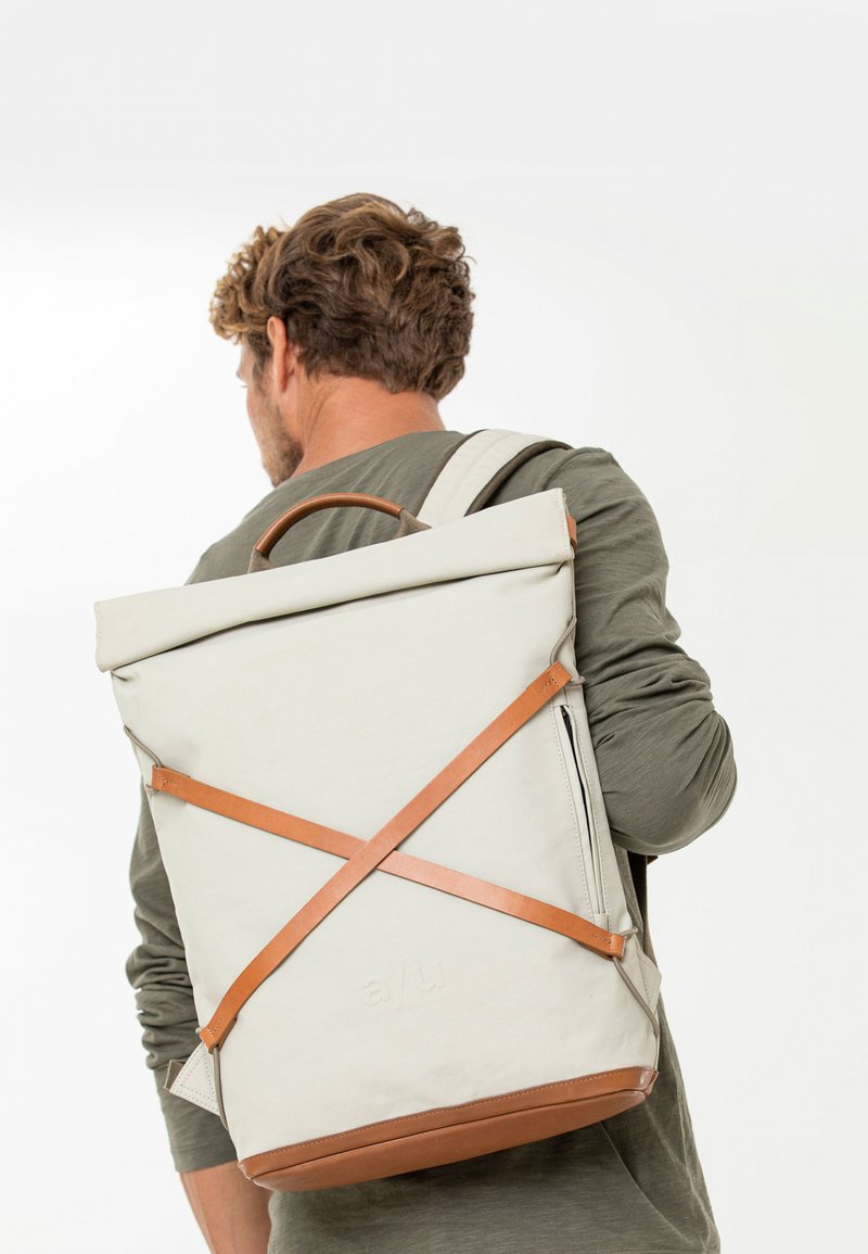 Zaino roll-top in tessuto beige chiaro con dettagli in pelle. Presenta spallacci regolabili, una maniglia superiore e una superficie liscia e durevole.