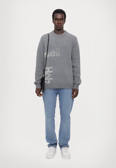 Grauer Pullover mit strukturiertem Schriftzug, kombiniert mit hellblauen Jeans und weißen Sneakers. Das Model trägt außerdem eine schwarze Umhängetasche.