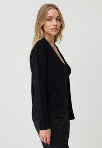 Cardigan nero con scollo a V profondo, maniche lunghe e un tessuto leggermente strutturato che aggiunge un leggero luccichio. Design aderente.