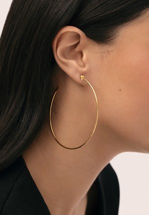 Primer plano de la oreja de una mujer con cabello largo y oscuro, que lleva un gran pendiente de aro delgado de oro y un pequeño pendiente de corazón de oro.