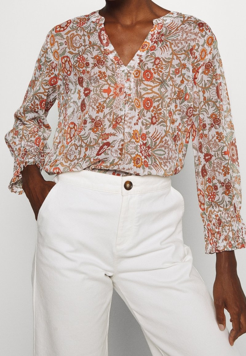 Blusa fluida de manga larga en tela ligera y transparente con un patrón floral en naranja, rojo y verde sobre un fondo claro; escote en V, puños con fruncido.