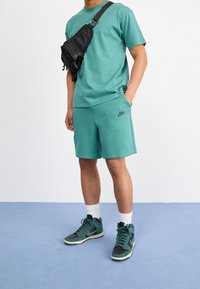 Turkusowa bawełniana koszulka i szorty z czarnym logo Nike. Czarna torba na ramię. Zielone i szare trampki wysokie. Białe skarpetki za kostkę.