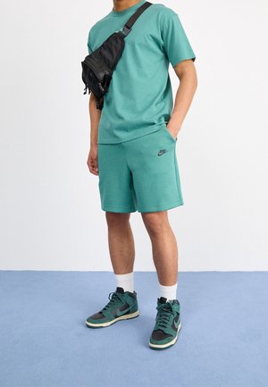 Turkoosi puuvillapaita ja shortsit, joissa on musta Nike-logo. Musta olkalaukku. Vihreät ja harmaat korkeavartiset lenkkarit. Valkoiset urheilusukat.