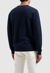 Marineblaues Sweatshirt mit langen Ärmeln, gerippten Bündchen und Saum. Glatte Textur, Rundhalsdesign, von hinten betrachtet. Getragen mit hellfarbigen Hosen.