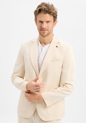 Homme aux cheveux châtain clair portant une veste de costume crème et une chemise blanche, ajustant la veste d'une main sur un fond blanc uni.