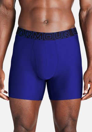 Männlicher Oberkörper trägt eng anliegende blaue Boxershorts mit schwarzem Bund und blauem "ARMOUR"-Schriftzug, steht vor weißem Hintergrund.