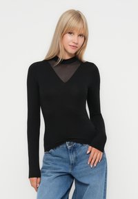 Black long-sleeve top with a sheer mesh V-neck detail and fitted design, paired with light blue high-waisted jeans. Smooth texture.