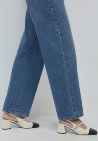 Chaussures à talons blocs crème et noires avec un design à bride arrière ; cuir texturé, bout arrondi et détail de sangle à la cheville, accompagnées de jeans en denim bleu.