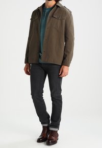 Olivgrüne, knöpfbare Jacke mit zwei Brusttaschen, die über einem türkisfarbenen Henley-Shirt getragen wird. Kombiniert mit dunklen Jeans und braunen Schnürstiefeln.