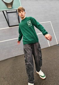 Grön texturerad tröja med vitt "S"-bokstav detaljer, matchad med grå cargobyxor med fickor och sneakers med gröna accenter.
