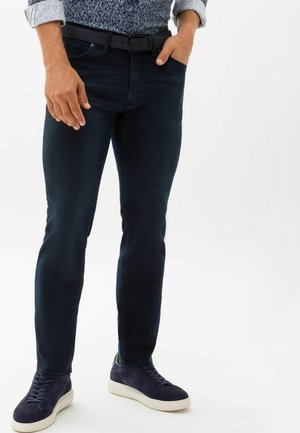 Dark blue slim-fit jeans with a smooth texture, featuring a classic five-pocket design, worn with greenish-blue suede sneakers.
