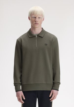 Junge Person mit hellem Haar, die einen olivgrünen Pullover mit Reißverschluss am Kragen und langen Ärmeln sowie schwarze Hosen trägt, steht vor einem schlichten Hintergrund.