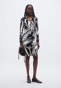 Svart-hvitt abstrakt kjole med virvelmønster, lange ermer, innsvingt midje og knelang lengde; kombinert med flate sko og en svart veske.