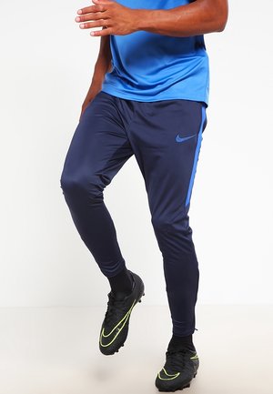 Personne portant un pantalon de sport bleu et un maillot bleu assorti, levant un genou, avec des crampons noirs ornés d'accents jaunes.