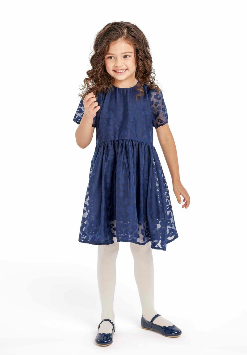 Robe bleu marine à manches courtes, en tissu texturé, avec une taille froncée. Des collants de couleur claire et des chaussures bleu marine assorties complètent la tenue.