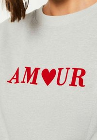 Sweat-shirt gris avec le mot "AMOUR" en lettres rouges, un cœur rouge remplaçant la lettre "O". Col rond classique, tissu en mélange de coton.