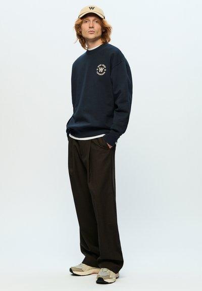 Sweatshirt marine avec logo rond, casquette beige, pantalon brun ample et baskets gris clair. Le tissu semble doux et décontracté.