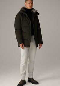 Donkergroene gewatteerde jas met een nepbont gevoerde kap, knoopsluiting aan de voorkant en twee zijzakken, gedragen met witte jeans en donkere laarzen.