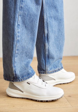 Sneakers bianche con lacci indossate con jeans di denim blu, in piedi su un pavimento in legno chiaro.