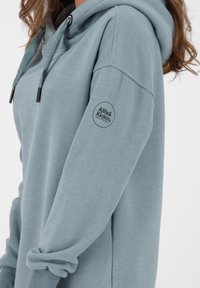 Vaaleansininen huppari rennolla istuvuudella, jossa on naruilla säädettävä huppu ja pyöreä logo vasemmassa hihassa. Valmistettu pehmeästä, teksturoidusta kankaasta.