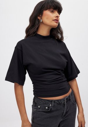 Schwarzes, kurzärmliges Top mit Stehkragen aus weichem Stoff, mit taillierter Silhouette und Raffung an der Taille. Kombiniert mit dunklen Jeans.