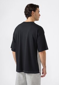 Hombre con pelo rizado oscuro que lleva una camisa de mangas cortas negra y gris holgada, y pantalones de chándal grises, de perfil a la derecha contra un fondo blanco.