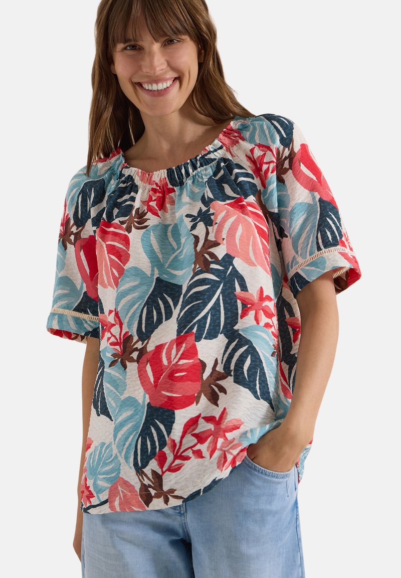 Femme souriante, portant un chemisier à manches courtes à motif floral coloré et un jean bleu clair, une main dans la poche, fond blanc.