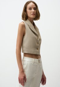 Beige verkürzte Weste mit maßgeschneidertem Design, ausgestattet mit einem Kragen und drei Knöpfen, kombiniert mit weißen hochgeschnittenen Hosen. Glatte Textur.