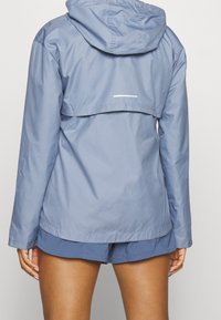 Persona con chaqueta con capucha de color azul claro y pantalones cortos de color azul oscuro, vista desde atrás sobre un fondo claro.