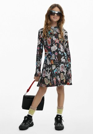 Langärmliges schwarzes Kleid mit bunten Cartoon-Tiermotiven, getragen mit pastellfarbenen Socken, schwarzen Sneakern und einer kleinen schwarzen Tasche mit rotem Riemen.