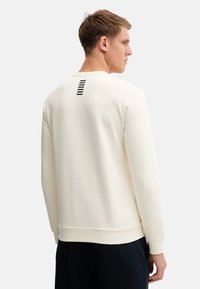 Felpa color crema con scollatura rotonda, maniche lunghe e un sottile dettaglio a righe sul retro. Tessuto morbido con una vestibilità comoda.