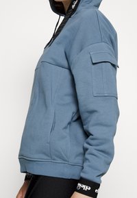 Blå hoodie med en framficka, sidsprund och långa ärmar. Har en justerbar huva och elastiska manschetter vid fållen och ärmarna.