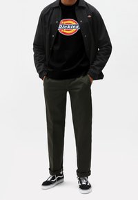 Person som bär svart jacka, svart Dickies-logotshirt, mörkgröna byxor och svartvita sneakers, med händerna i fickorna, står framför en vit bakgrund.