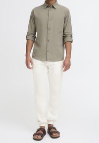 Homme portant une chemise boutonnée vert olive clair avec manches retroussées, pantalon de jogging blanc et sandales marron à double lanière, debout devant un fond blanc.