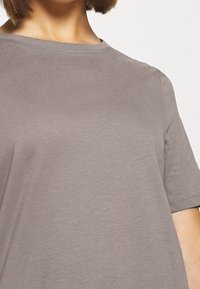 Persona che indossa una t-shirt di cotone beige, a maniche corte e con scollatura rotonda, su uno sfondo bianco.
