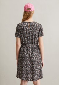 Floral jurk van zwarte stof met korte mouwen, met een getailleerde taille en een sleuteldetail aan de achterkant. Gecombineerd met een roze pet.
