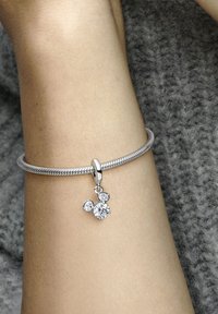 Srebrna zapestnica z pletenim dizajnom, ki ima viseč čar s tremi prozornimi dragimi kamni različnih velikosti. Teksturo ima sivo ozadje.