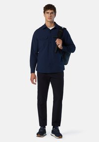 Marineblaue Langarmhemd mit zwei Brusttaschen, kombiniert mit marineblauen Hosen und schwarzen Sneakern. Das Modell trägt einen schwarzen Rucksack.