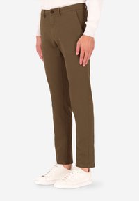 Pantaloni chinos marroni realizzati in tessuto di cotone resistente, vestibilità slim, dotati di tasche frontali, chiusura con bottoni e dettagli di cuciture a contrasto. Indossati con scarpe da ginnastica bianche.