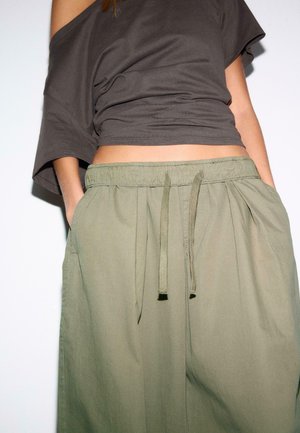 Ženska oblečena v olivno zelene hlače z vrvico in ohlapen temno rjav top z odkrito ramo, z rokami v žepih, obrezana pri sredini trebuha.