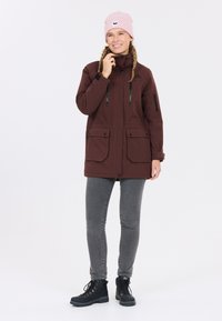 Bruine parka jas met dubbele voorzakken, ritsaccenten en een verstelbare capuchon, gecombineerd met grijze jeans en zwarte enkellaarsjes. Roze beanie gedragen.