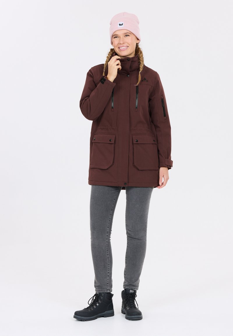 Bruine parka jas met dubbele voorzakken, ritsaccenten en een verstelbare capuchon, gecombineerd met grijze jeans en zwarte enkellaarsjes. Roze beanie gedragen.