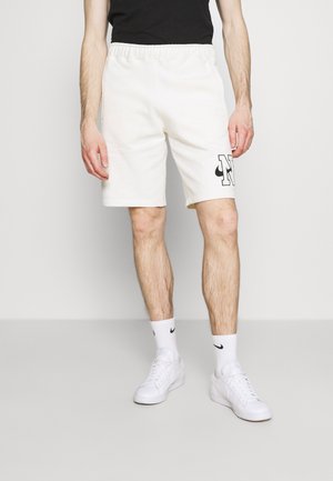 Mann trägt weiße Nike-Shorts mit schwarzem Logo, weiße Nike-Socken, weiße Sneakers und ein schwarzes Shirt, steht vor einem einfarbigen Hintergrund.
