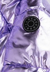 Błyszcząca metaliczna purpurowa tkanina pikowana z okrągłą czarno-białą łatą z napisem "Cocodrillo" na powierzchni.
