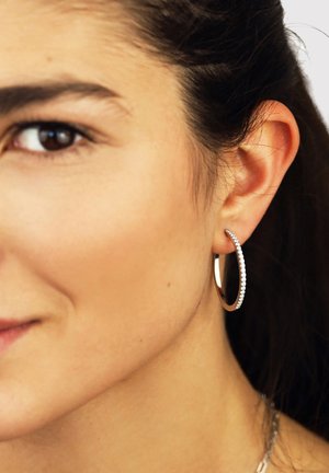 Nahaufnahme eines Gesichts einer Frau mit einem silbernen Creolenohrring, der kleine eingearbeitete Steine aufweist, dunklem, zurückgebundenem Haar und braunen Augen.