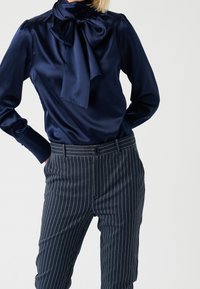 Blusa di raso blu navy con un grande colletto a fiocco, abbinata a pantaloni blu navy a righe con vita standard e chiusura a bottone.